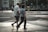 An elderly couple walks hand in hand across a city street. The urban environment features buildings, parked bicycles, and blurred figures in the background. The scene captures a moment of quiet companionship amidst a busy setting.
