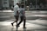 An elderly couple walks hand in hand across a city street. The urban environment features buildings, parked bicycles, and blurred figures in the background. The scene captures a moment of quiet companionship amidst a busy setting.