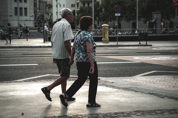 An elderly couple walks hand in hand across a city street. The urban environment features buildings, parked bicycles, and blurred figures in the background. The scene captures a moment of quiet companionship amidst a busy setting.