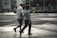 An elderly couple walks hand in hand across a city street. The urban environment features buildings, parked bicycles, and blurred figures in the background. The scene captures a moment of quiet companionship amidst a busy setting.