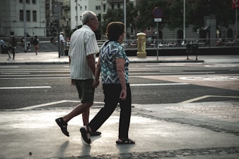 An elderly couple walks hand in hand across a city street. The urban environment features buildings, parked bicycles, and blurred figures in the background. The scene captures a moment of quiet companionship amidst a busy setting.