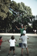 A person is creating large soap bubbles using a rope apparatus, surrounded by three children eagerly reaching out to interact with the bubbles. The setting is an outdoor area with trees in the background, conveying a playful and lively atmosphere.