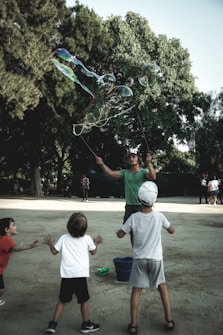 A person is creating large soap bubbles using a rope apparatus, surrounded by three children eagerly reaching out to interact with the bubbles. The setting is an outdoor area with trees in the background, conveying a playful and lively atmosphere.