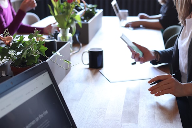 A group of people are working at a wooden table, some using laptops and others on their phones. Potted plants and mugs are placed on the table, contributing to a relaxed office setting. Natural light illuminates the scene, emphasizing a casual yet professional atmosphere.