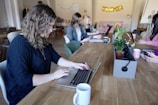 A group of people working at a long wooden table in a cozy office space. Several laptops are open, and coffee mugs are scattered across the table. Plants and a flower arrangement add a touch of greenery. The focus is on a person with curly hair typing on a laptop in the foreground.