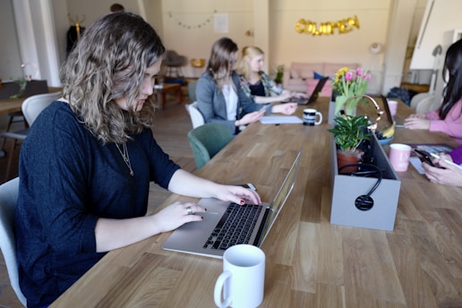 A group of people working at a long wooden table in a cozy office space. Several laptops are open, and coffee mugs are scattered across the table. Plants and a flower arrangement add a touch of greenery. The focus is on a person with curly hair typing on a laptop in the foreground.
