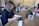 A group of people working at a long wooden table in a cozy office space. Several laptops are open, and coffee mugs are scattered across the table. Plants and a flower arrangement add a touch of greenery. The focus is on a person with curly hair typing on a laptop in the foreground.