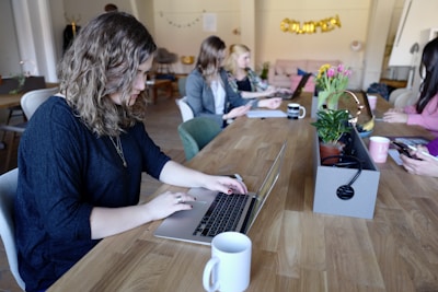 A group of people working at a long wooden table in a cozy office space. Several laptops are open, and coffee mugs are scattered across the table. Plants and a flower arrangement add a touch of greenery. The focus is on a person with curly hair typing on a laptop in the foreground.