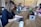 A group of people working at a long wooden table in a cozy office space. Several laptops are open, and coffee mugs are scattered across the table. Plants and a flower arrangement add a touch of greenery. The focus is on a person with curly hair typing on a laptop in the foreground.
