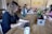 A group of people working at a long wooden table in a cozy office space. Several laptops are open, and coffee mugs are scattered across the table. Plants and a flower arrangement add a touch of greenery. The focus is on a person with curly hair typing on a laptop in the foreground.