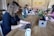 A group of people working at a long wooden table in a cozy office space. Several laptops are open, and coffee mugs are scattered across the table. Plants and a flower arrangement add a touch of greenery. The focus is on a person with curly hair typing on a laptop in the foreground.