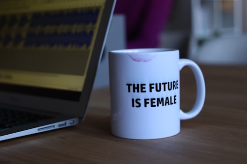 A white ceramic mug featuring the slogan 'THE FUTURE IS FEMALE' is placed on a wooden table. There is a visible lipstick mark on the rim of the mug. Beside the mug, part of an open laptop is visible, displaying what appears to be soundwave patterns on the screen.