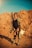 A person stands in a desert-like environment, wearing a rugged outfit with goggles perched on their head and tribal face paint. They hold a large gun in one hand and a bleached animal skull in the other. The background consists of rocky, barren terrain under a clear blue sky.