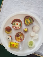 A white plate with a variety of Indian desserts and snacks. There are terracotta cups filled with custard and garnished with fresh fruit slices like kiwi, pomegranate seeds, and strawberries. A gulab jamun sits in a small dish with syrup. Served alongside are slices of white radish and cucumber. There is also a piece of silver-coated sweet and a yellow, spongy dessert garnished with red and green toppings. A white spoon is placed on the plate.