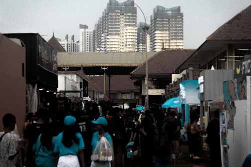 A bustling street market is filled with people walking and browsing booths. The setting is surrounded by modern high-rise buildings in the background, while the market structures have traditional, tiled roofs. Many of the attendees are wearing casual clothing, with some wearing uniforms or promotional shirts.