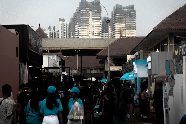A bustling street market is filled with people walking and browsing booths. The setting is surrounded by modern high-rise buildings in the background, while the market structures have traditional, tiled roofs. Many of the attendees are wearing casual clothing, with some wearing uniforms or promotional shirts.