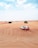 A white and red SUV is driving across sandy dunes under a blue sky with scattered clouds. The expansive desert landscape features gentle slopes and sparse vegetation, conveying a sense of adventure and isolation.