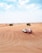A white and red SUV is driving across sandy dunes under a blue sky with scattered clouds. The expansive desert landscape features gentle slopes and sparse vegetation, conveying a sense of adventure and isolation.