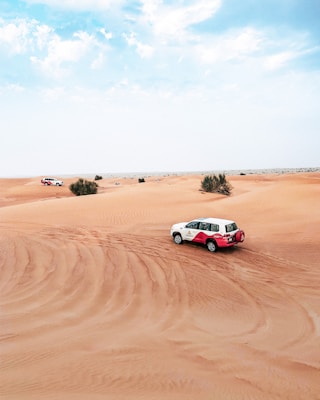 A white and red SUV is driving across sandy dunes under a blue sky with scattered clouds. The expansive desert landscape features gentle slopes and sparse vegetation, conveying a sense of adventure and isolation.
