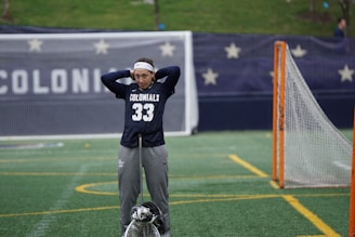 A person wearing a navy blue 'COLONIALS' sports jersey with the number 33 stands on a sports field, holding a lacrosse stick and wearing a headband. In the background, there is a banner with stars and the word 'COLONIALS' visible. The field has green artificial turf with yellow lines and there is a lacrosse goal to the side.