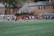 A group of people, primarily young athletes, are gathered on a sports field, some dressed in uniforms consisting of navy blue and red colors. In the background, there are several red-brick buildings and blossoming trees, suggesting a springtime setting. A goalpost can be seen, indicating that this is likely a game of soccer or lacrosse. The scene is lively and bustling with activity.