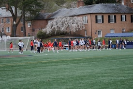 A group of people, primarily young athletes, are gathered on a sports field, some dressed in uniforms consisting of navy blue and red colors. In the background, there are several red-brick buildings and blossoming trees, suggesting a springtime setting. A goalpost can be seen, indicating that this is likely a game of soccer or lacrosse. The scene is lively and bustling with activity.