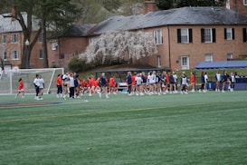 A group of people, primarily young athletes, are gathered on a sports field, some dressed in uniforms consisting of navy blue and red colors. In the background, there are several red-brick buildings and blossoming trees, suggesting a springtime setting. A goalpost can be seen, indicating that this is likely a game of soccer or lacrosse. The scene is lively and bustling with activity.