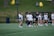 A group of female athletes in sportswear are gathered on a green field, possibly preparing or practicing for a game. They are wearing white and navy uniforms, some with numbers on the back. In the background, there are outdoor chairs on a grassy hill and a bench area with a roof structure, suggesting a sports facility. Small yellow balls are scattered on the turf.