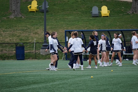 A group of female athletes in sportswear are gathered on a green field, possibly preparing or practicing for a game. They are wearing white and navy uniforms, some with numbers on the back. In the background, there are outdoor chairs on a grassy hill and a bench area with a roof structure, suggesting a sports facility. Small yellow balls are scattered on the turf.