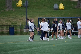 A group of female athletes in sportswear are gathered on a green field, possibly preparing or practicing for a game. They are wearing white and navy uniforms, some with numbers on the back. In the background, there are outdoor chairs on a grassy hill and a bench area with a roof structure, suggesting a sports facility. Small yellow balls are scattered on the turf.
