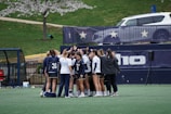 A group of female athletes in sports uniforms huddling on a grassy field, surrounded by a fence with star decorations and a white vehicle in the background. The athletes are wearing matching jerseys with numbers and the environment suggests a sporting event preparation.