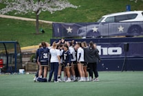 A group of female athletes in sports uniforms huddling on a grassy field, surrounded by a fence with star decorations and a white vehicle in the background. The athletes are wearing matching jerseys with numbers and the environment suggests a sporting event preparation.