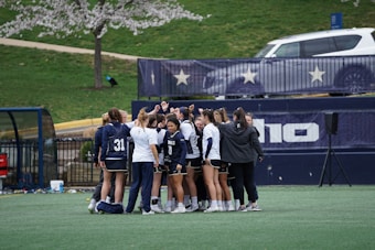 A group of female athletes in sports uniforms huddling on a grassy field, surrounded by a fence with star decorations and a white vehicle in the background. The athletes are wearing matching jerseys with numbers and the environment suggests a sporting event preparation.