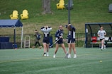 Three female lacrosse players are standing on a field, adjusting their equipment and preparing for play. They wear navy blue uniforms with white numbers. In the background, bright yellow and gray chairs and a blue tent are visible, along with a few people walking.