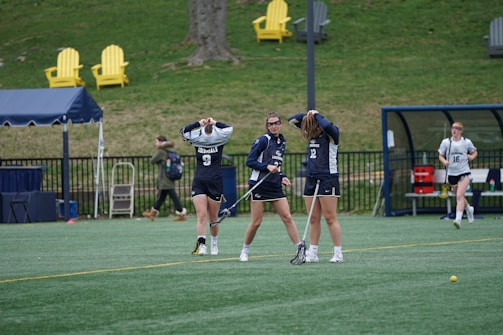 Three female lacrosse players are standing on a field, adjusting their equipment and preparing for play. They wear navy blue uniforms with white numbers. In the background, bright yellow and gray chairs and a blue tent are visible, along with a few people walking.