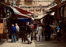A bustling outdoor market with people walking through a narrow alley lined with shops. The stalls display a variety of goods including textiles and crafts under colorful awnings, with a historical architectural backdrop.