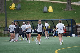 Several female athletes wearing sports uniforms and protective eyewear are gathered on a lacrosse field. They are holding lacrosse sticks and appear to be either warming up or discussing strategy. In the background, there are benches and a few colorful outdoor chairs on a grassy area.