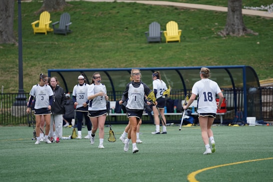 Several female athletes wearing sports uniforms and protective eyewear are gathered on a lacrosse field. They are holding lacrosse sticks and appear to be either warming up or discussing strategy. In the background, there are benches and a few colorful outdoor chairs on a grassy area.