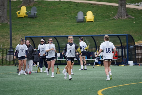 Several female athletes wearing sports uniforms and protective eyewear are gathered on a lacrosse field. They are holding lacrosse sticks and appear to be either warming up or discussing strategy. In the background, there are benches and a few colorful outdoor chairs on a grassy area.