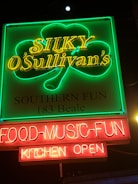 A vibrant neon sign displays the text 'Silky O'Sullivan's' in yellow neon with a green shamrock outline around it. Below, the words 'Southern Fun' and '183 Beale' are illuminated. At the bottom, red neon signs display 'FOOD-MUSIC-FUN' and 'KITCHEN OPEN'. The setting appears to be a nighttime urban environment.