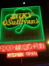 A vibrant neon sign displays the text 'Silky O'Sullivan's' in yellow neon with a green shamrock outline around it. Below, the words 'Southern Fun' and '183 Beale' are illuminated. At the bottom, red neon signs display 'FOOD-MUSIC-FUN' and 'KITCHEN OPEN'. The setting appears to be a nighttime urban environment.