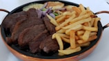 A hearty meal featuring a large serving of grilled steak accompanied by a generous portion of golden French fries. Red onion slices and zucchini are also visible on the plate, providing a burst of color. The food is served in a dark, circular pan with copper-colored handles.