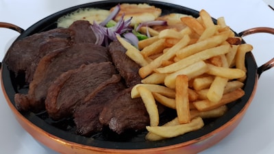 A hearty meal featuring a large serving of grilled steak accompanied by a generous portion of golden French fries. Red onion slices and zucchini are also visible on the plate, providing a burst of color. The food is served in a dark, circular pan with copper-colored handles.