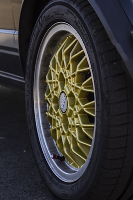 A close-up view of a car tire with a yellow BBS alloy wheel, featuring intricate spoke patterns. The tire is mounted on a car, and the brand name BBS is visible at the center of the wheel hub.