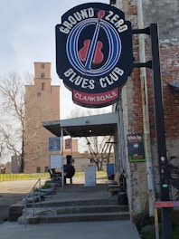 A sign for the Ground Zero Blues Club in Clarksdale is prominently displayed above the entrance. The exterior of the building has a rustic and weathered look, with exposed brick and a tin roof overhanging the entrance. There are posters and signs on the wall adjacent to the entrance. Outdoor seating and a charcoal smoker can be seen on the patio. In the background, there is an old tower and leafless trees.