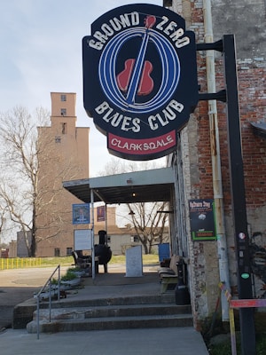 A sign for the Ground Zero Blues Club in Clarksdale is prominently displayed above the entrance. The exterior of the building has a rustic and weathered look, with exposed brick and a tin roof overhanging the entrance. There are posters and signs on the wall adjacent to the entrance. Outdoor seating and a charcoal smoker can be seen on the patio. In the background, there is an old tower and leafless trees.