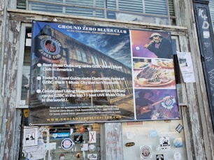 A poster for the Ground Zero Blues Club is displayed on a weathered wooden exterior wall. The poster features images of the club, a male singer with a microphone, a plate of food including ribs and cornbread, and a guitarist playing an electric guitar. Text highlights the club's rankings and accolades from various publications as a top blues music destination in Clarksdale, Mississippi.
