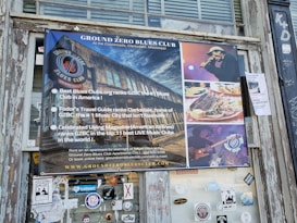 A poster for the Ground Zero Blues Club is displayed on a weathered wooden exterior wall. The poster features images of the club, a male singer with a microphone, a plate of food including ribs and cornbread, and a guitarist playing an electric guitar. Text highlights the club's rankings and accolades from various publications as a top blues music destination in Clarksdale, Mississippi.