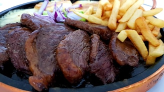 A plate filled with grilled meat slices, crispy golden french fries, slices of red onions, and some green vegetables. The food is attractively arranged and looks appetizing.