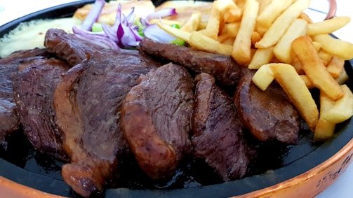 A plate filled with grilled meat slices, crispy golden french fries, slices of red onions, and some green vegetables. The food is attractively arranged and looks appetizing.
